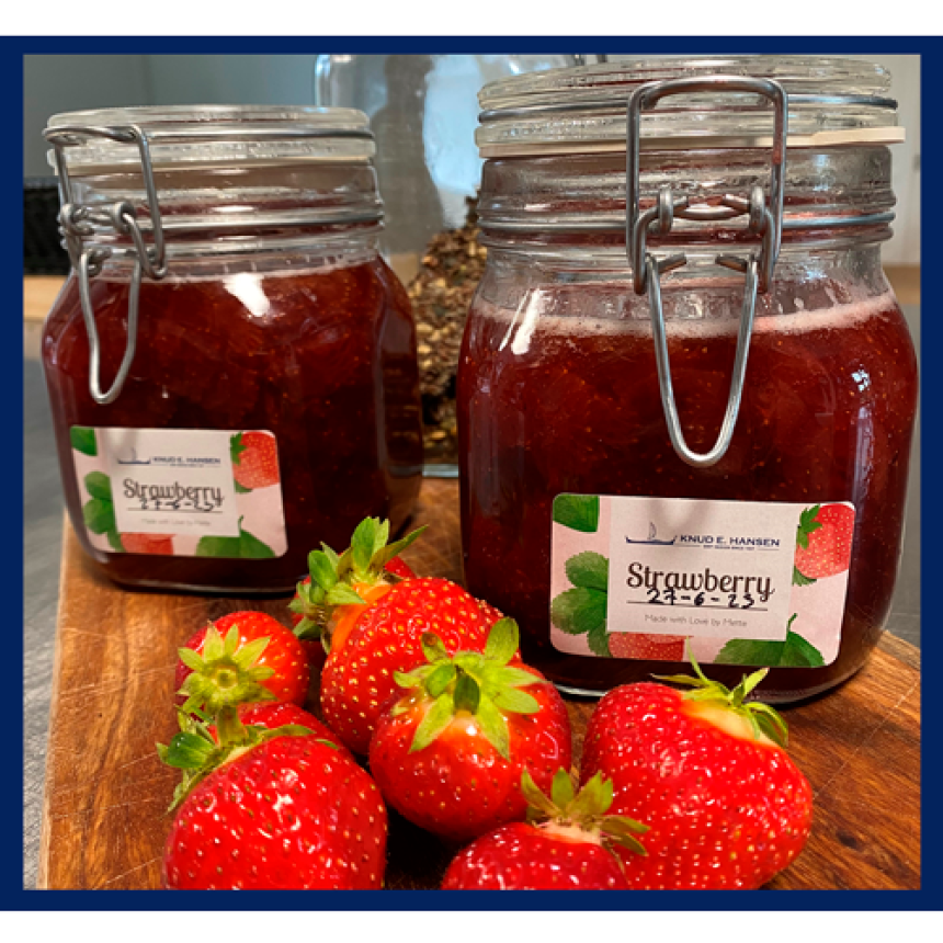 Syltetøjsglas med jordbærmarmelade og en etiket med teksten Strawberry. Står på et træbræt, omringet af jordbær.