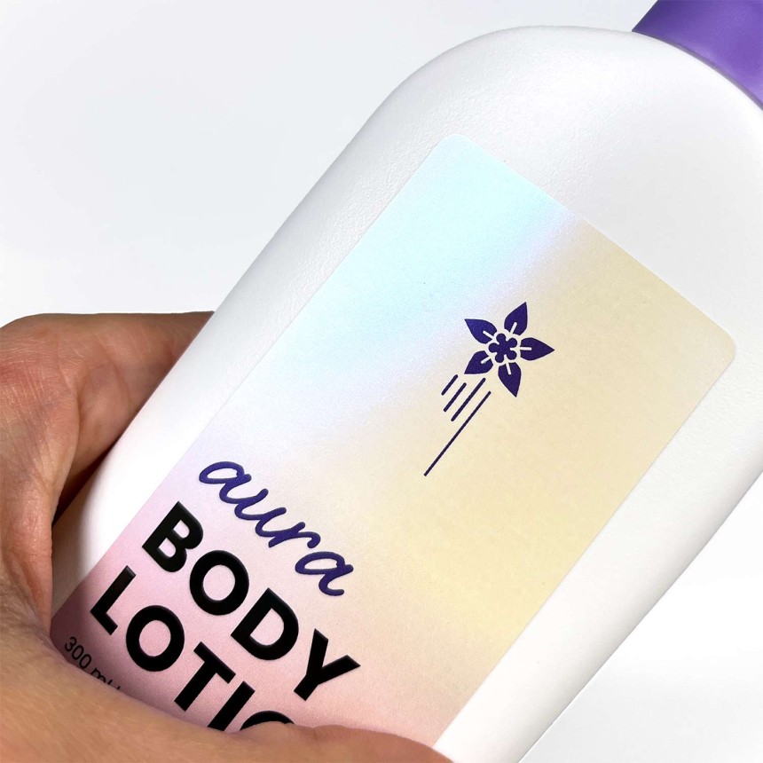 Etiket i perslemorsskinnende folie med teksten "aura body lotion". Her ses den skinnende effekt på en hvid flaske.