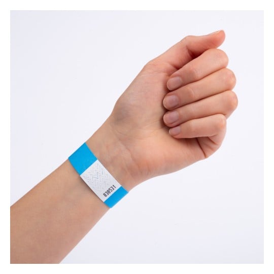 Billedet viser en hånd iført et blå- og hvidstribet armbånd. Armbåndet har en kode trykt på det, som muligvis angiver adgang eller entré til en begivenhed.