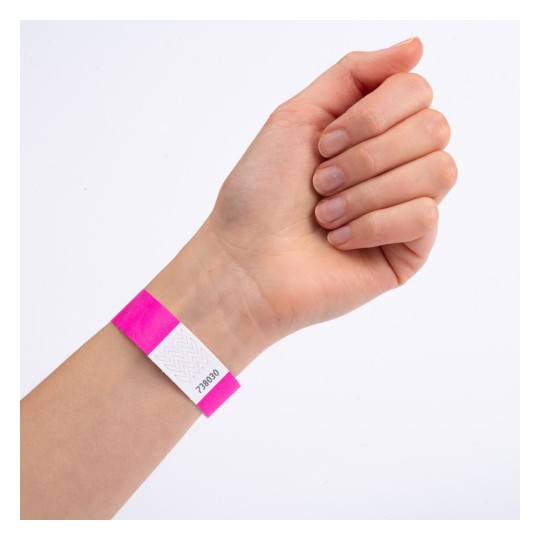 Billedet viser en hånd med et pink armbånd, der har en hvid etiket og et nummer trykt på. Hånden er placeret med fingrene lukkede, og armbåndet er tydeligt synligt.