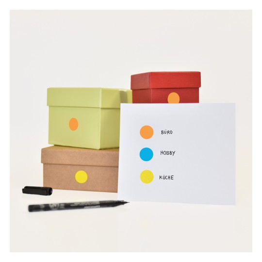 Billedet viser flere farverige kasser stablet sammen med et kort, der har tre mærkede prikker: orange for 'BÜRO', blå for 'HOBBY' og gul for 'KÜCHE'. En sort pen er også synlig ved siden af kasserne.
