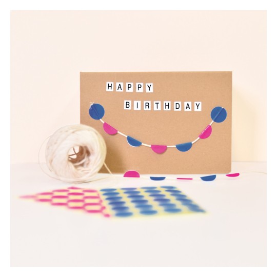 Billedet viser et fødselsdagskort med ordene 'HAPPY BIRTHDAY' på. Det har en dekorativ snor af blå og lyserøde cirkler fastgjort til kortet. I nærheden er der en rulle snor og et mønstret papir med farverige prikker.