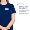 Billedet viser en person iført en marineblå skjorte med et navneskilt, der lyder 'B. Carlos, Marketing Director.' Der er også en beskrivelse, der angiver, at navneskiltet er selvklæbende og kan sidde på tøj pålideligt uden at efterlade rester.