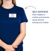 Billedet viser en person iført en blå skjorte med et navneskilt, der lyder 'B. Carlos, Marketing Director'. Der er tekst, der angiver den selvklæbende natur af skiltet, som kan klæbe til tøj og nemt fjernes uden at efterlade rester.