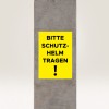 Billedet viser et gult skilt på en betonpille, hvor der står 'BITTE SCHUTZ-HELM TRAGEN!', hvilket oversættes til 'Venligst brug hjelm!' og angiver et sikkerhedskrav.