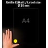 Billedet viser en hånd placeret ved siden af et A4-ark. En cirkulær gul label med en diameter på 30 mm er placeret i det nederste hjørne af papiret. Baggrunden er mørk, hvilket forbedrer synligheden.